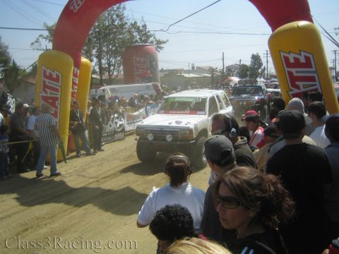 1990 Ford Bronco - white off-road SUV crosses a dusty start area under an inflatable Tecate arch with a dense crowd of onlookers.