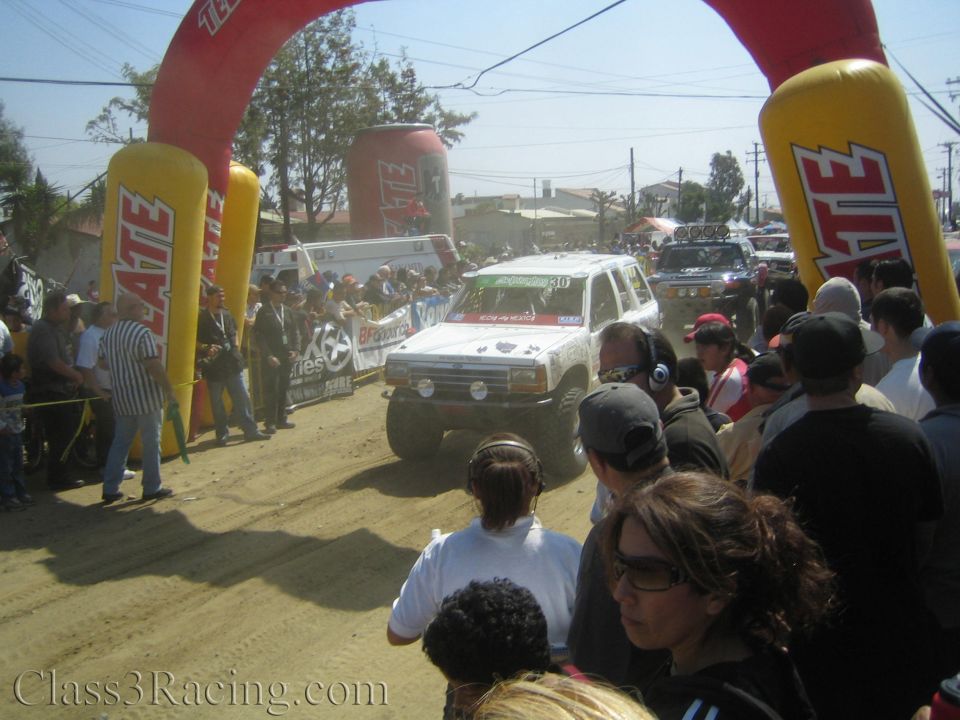 White off-road SUV crossing a dusty start line under a Tecate arch