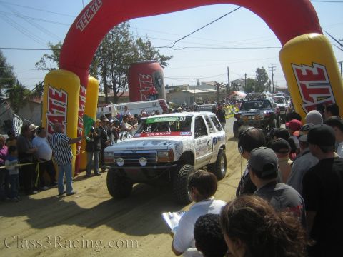 1990 Ford Bronco - white Ford SUV racer numbered 30 sits at a dusty dirt start line under a red inflatable Tecate arch with crowds nearby.