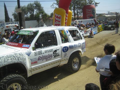 1990 Ford Bronco - white off-road race SUV lettered Chupakabras Racing and numbered 301 kicks up dust beside the crowd at the Tecate start area.