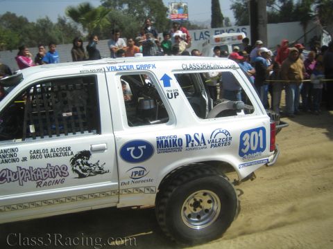 1990 Ford Bronco - Side profile of a white Chupakabras Racing off-road SUV numbered 301 covered in sponsor decals on a dirt course before a crowd.