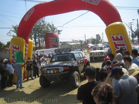 1990 Ford Bronco - black off-road race truck with roof-mounted lights idles under the large red Tecate inflatable arch as a flagger and crowd look on.