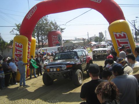 1990 Ford Bronco - Toyota FJ Cruiser off-road race vehicle staged under a red and yellow Tecate inflatable arch at a desert race start, surrounded by spectato…