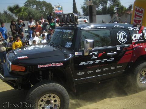 1990 Ford Bronco - Side profile of a black Toyota-badged off-road race truck numbered 302 with Nitto and KC decals and a roof basket on a dirt course.