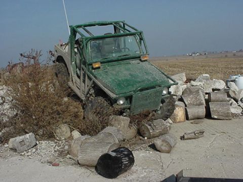 1992 Ford Bronco - A green off-road vehicle is partially submerged in brush and rocks, with a clear sky in the background.