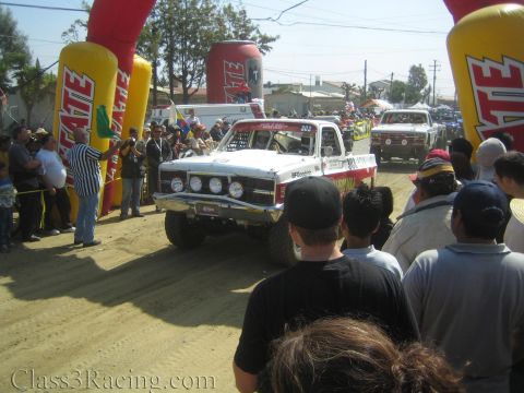 1990 Ford Bronco - white off-road race truck with stacked headlights waits at the Tecate start arch as a flagger raises a green flag before the crowd.