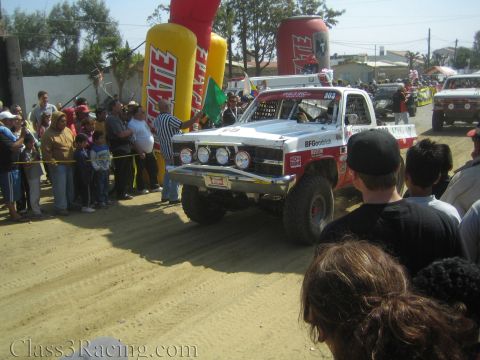 1990 Ford Bronco - white off-road race truck numbered 300 with BFGoodrich decals pulls away from the Tecate start arch onto the dirt course.
