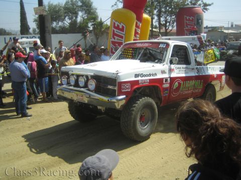 1990 Ford Bronco - red-and-white off-road race truck numbered 303 lettered California Tomatoes and BFGoodrich leaves the Tecate start arch.