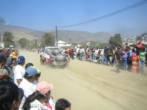 1990 Ford Bronco - off-road race truck speeds along a dusty desert road flanked by crowds, with arid hills in the background.