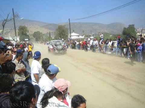1990 Ford Bronco - off-road race truck trails dust down a wide desert road lined with spectators beneath dry hills.