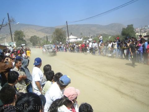 1990 Ford Bronco - white off-road race truck raises a heavy cloud of dust on a desert road packed with spectators below arid hills.