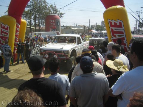 1990 Ford Bronco - white off-road race truck numbered 343 pulls from the Tecate start arch into the dust, viewed over a packed roadside crowd.