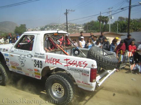 1990 Ford Bronco - White tube-frame off-road race truck number 349 reading Anger Issues kicks up dust on a dirt course before a hillside crowd.