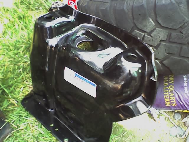 Black metal automotive part with a white sticker, resting on grass next to a tire and bag of garden soil