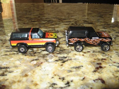 1986 Ford Bronco - Two toy Ford Broncos are displayed on a speckled countertop, one a sunset stripe design and the other adorned with flame graphics.