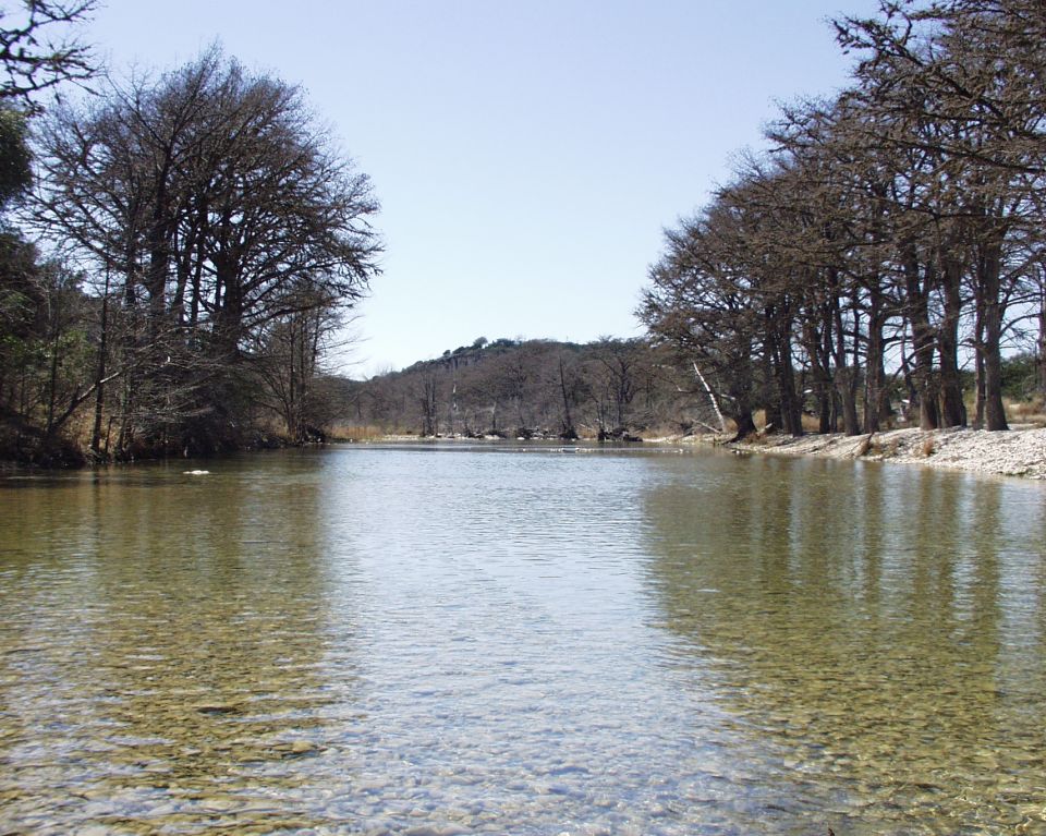 River with clear shallow water and rocky bottom, lined by bare trees under a clear blue sky