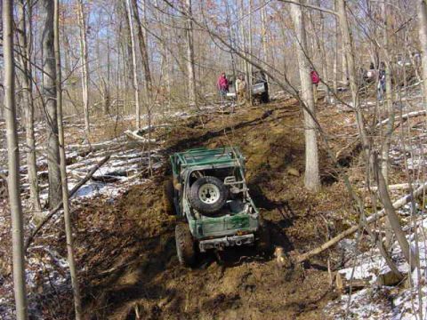 1992 Ford Bronco - An off-road vehicle navigates a steep, muddy incline in a wooded area with other vehicles visible further up the trail.