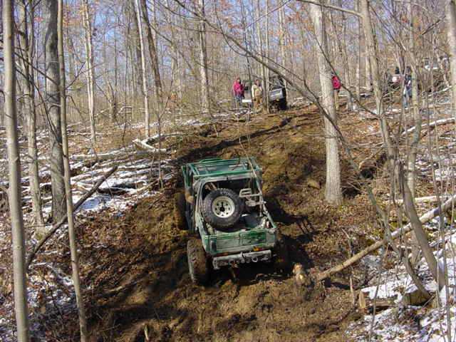 Green off-road vehicle climbing a muddy hill with other vehicles and people in the background