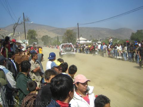 1990 Ford Bronco - white off-road race truck drives away down a dusty desert road framed by hills and a dense crowd of spectators.