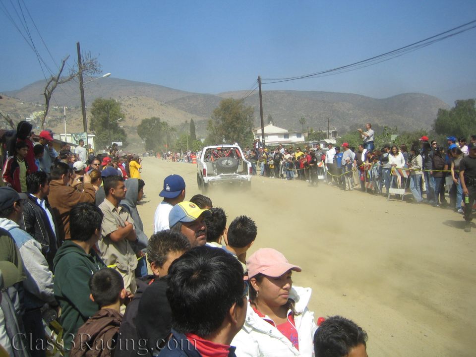 White race truck driving away in distance on dusty road past large crowd