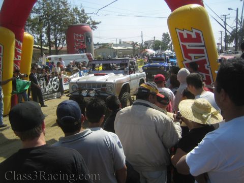 1990 Ford Bronco - vintage white off-road Bronco-style race truck waits at a start line beneath red Tecate inflatable arches amid a crowd.