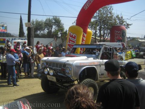 1990 Ford Bronco - white early-Bronco off-road race truck marked West Coast Broncos with a front light bar passes under a Tecate arch.