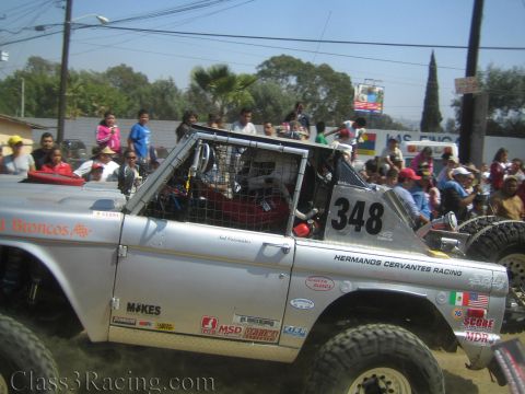 1990 Ford Bronco - Side view of a silver tube-frame off-road race truck number 348 from Hermanos Cervantes Racing with spectators behind.