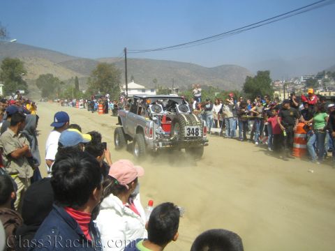 1990 Ford Bronco - Silver off-road race truck number 348 drives down a dusty desert road flanked by crowds and orange cones.