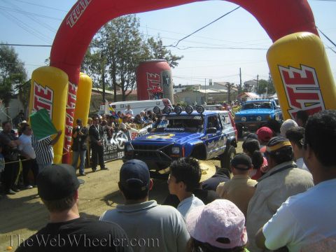 1990 Ford Bronco - blue Panasonic-liveried off-road race truck waits under a red and yellow inflatable Tecate start arch surrounded by spectators.