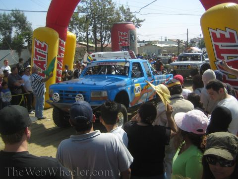 1990 Ford Bronco - blue Ford off-road race truck with NAPA decals stages near inflatable Tecate markers as spectators watch.