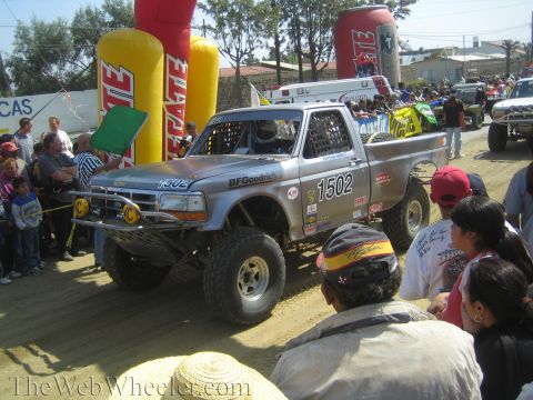 1990 Ford Bronco - silver Ford off-road race truck numbered 1502 with BFGoodrich decals stages on a dirt course as a flagger and crowd look on.