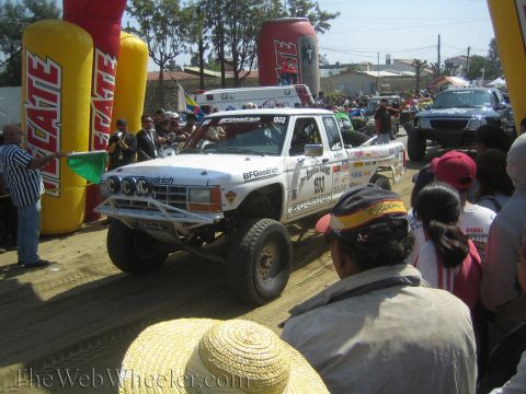 1990 Ford Bronco - white off-road race truck numbered 803 with a bumper light bar and BFGoodrich decals stages near a green flag and crowd.