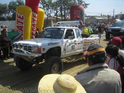 1990 Ford Bronco - white off-road race truck numbered 1503 with BFGoodrich and Camburg decals stages on the dirt beside an inflatable Tecate arch.