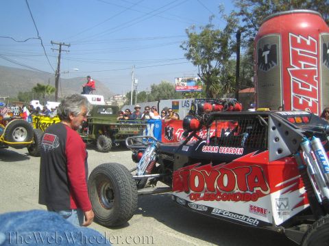 1990 Ford Bronco - red and black off-road race buggy lettered Toyota of Escondido and Sarah Bearden sits in a staging area beside a man holding a spare tire.
