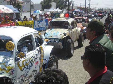 1990 Ford Bronco - Race-prepped Volkswagen Beetle off-road cars covered in sponsor decals queue at a street staging area as crowds watch.