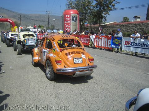 1990 Ford Bronco - orange number 411 Volkswagen Beetle off-road racer rolls past banners and crowds, with a giant Tecate can display behind.