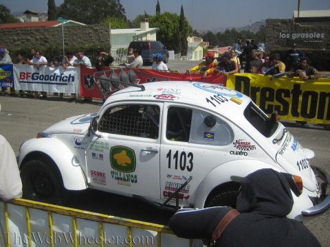 1990 Ford Bronco - white number 1103 Volkswagen Beetle off-road racer covered in decals sits behind a yellow barrier as a hooded spectator photographs it.