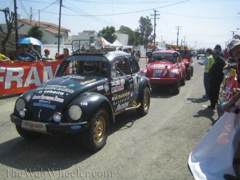 1990 Ford Bronco - black number 1148 Volkswagen Beetle off-road racer leads a red one and other vehicles past banners and onlookers.