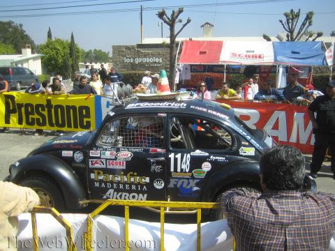 1990 Ford Bronco - black number 1148 Volkswagen Beetle off-road racer with Pacific and Akron decals sits behind a barrier as crowds gather.