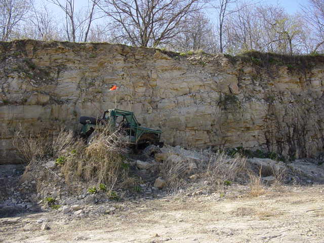 Green off-road vehicle with a roll cage and an orange flag on top, parked on a rocky incline next to a cliff