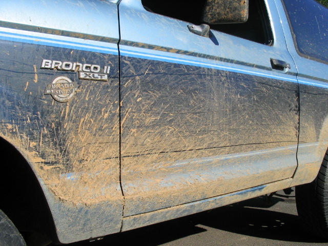 Side view of a blue Ford Bronco II covered in mud. It has a 'Bronco II XLT' badge and a 'Mountain States' emblem