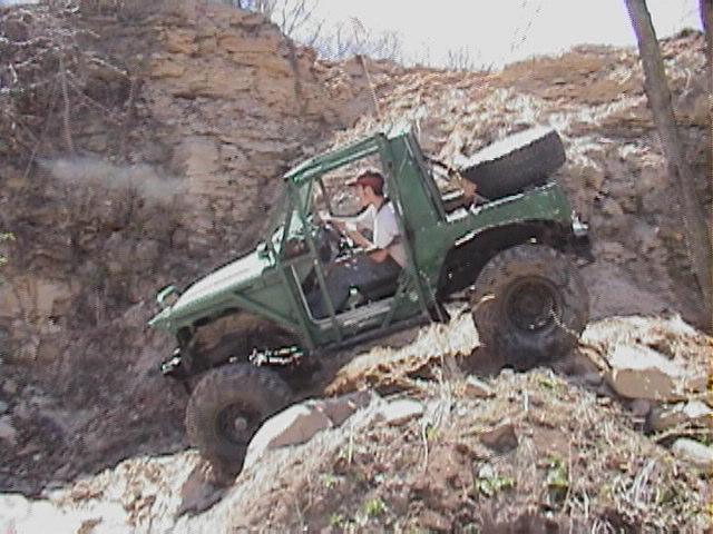 Green off-road vehicle with a spare tire on back climbing a steep, rocky hill. Driver is wearing a red cap