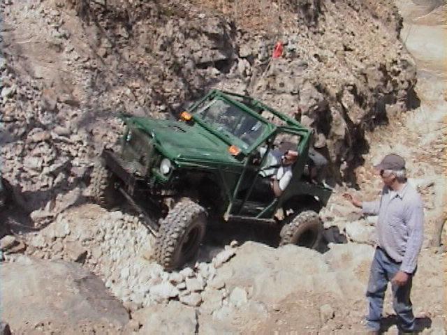 Green off-road vehicle with roll cage navigating rocky terrain, driver visible inside, man standing nearby