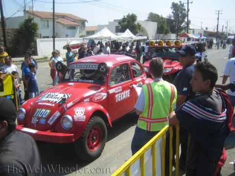 1990 Ford Bronco - red Tecate-sponsored number 1100 Volkswagen Beetle off-road racer sits at a street staging area beside an official in a safety vest.