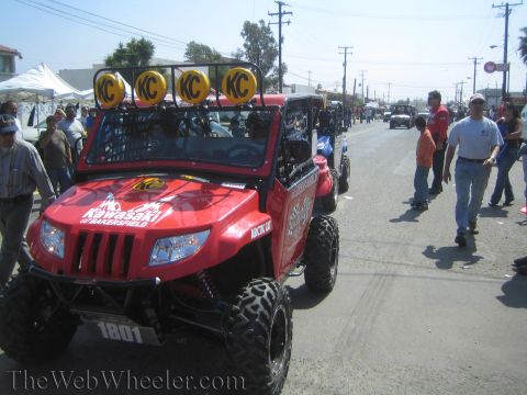 1990 Ford Bronco - red Arctic Cat side-by-side off-road UTV number 1801 with a KC roof light bar is parked on a sunny street with people walking by.