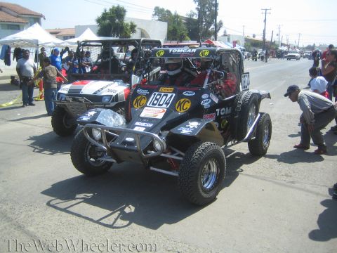 1990 Ford Bronco - black tube-frame off-road race buggy number 1804 with KC lights and Tomcar livery sits staged on a street with onlookers.