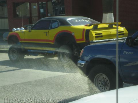 1990 Ford Bronco - yellow and red custom muscle-style coupe lifted on large off-road tires with a roof light bar sits next to a blue pickup truck.