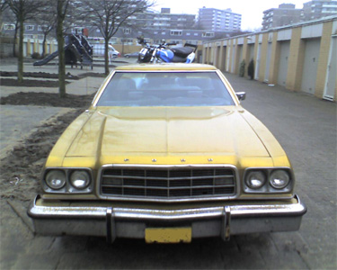 1973 Ford Ranchero - A yellow Ford sedan is parked in front of a row of garages, with a playground in the background.
