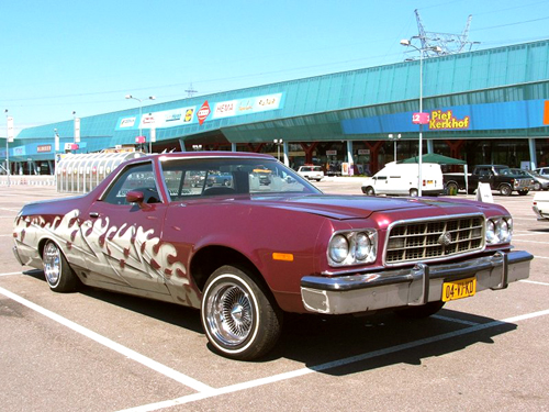 1973 Ford Ranchero with custom flame graphics on the side, parked in a lot. Yellow license plate visible