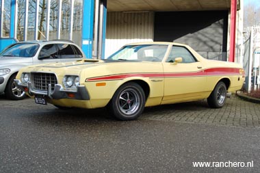 1973 Ford Ranchero - A yellow 1973 Ford Ranchero with a red stripe is parked outside a building.